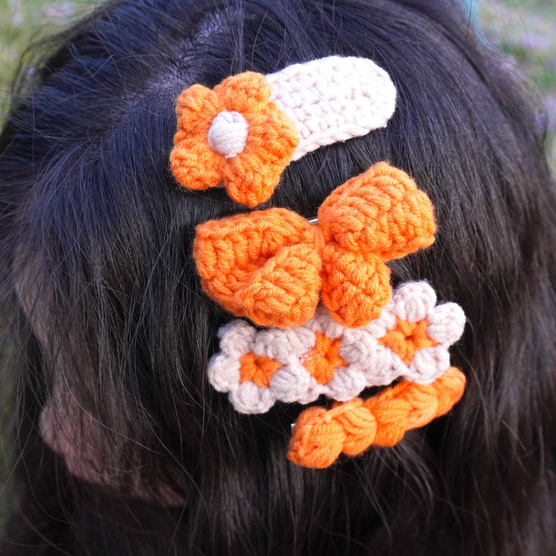 May include: A set of three crocheted hair clips in orange and beige. The clips feature a flower, a bow, and a more intricate design.