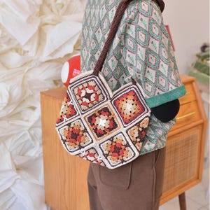 May include: A handmade crochet bag with a brown strap and a patchwork design. The bag features squares in shades of brown, orange, and cream, creating a geometric pattern. The bag is carried over the shoulder.