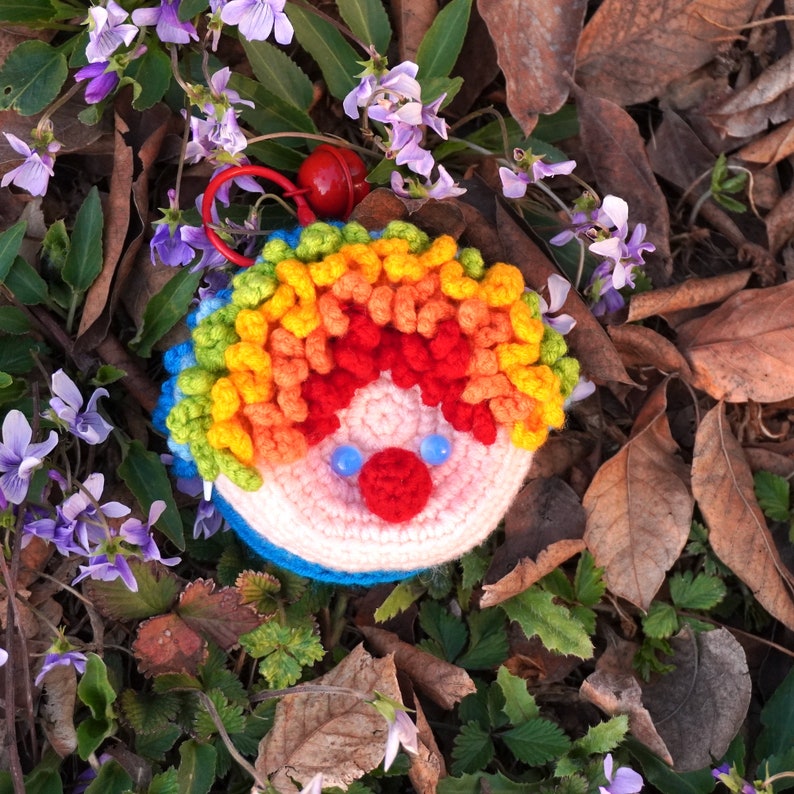 May include: A crocheted rainbow-colored clown face keychain with blue eyes and a red nose. The keychain is lying on a bed of purple flowers and brown leaves.