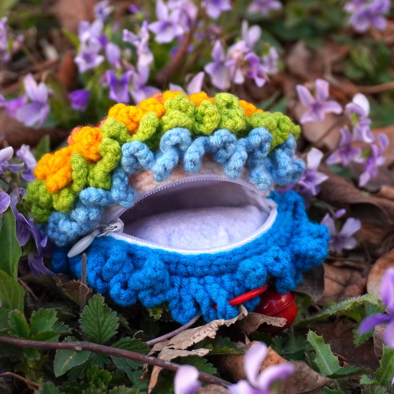 May include: A crocheted coin purse with a rainbow design and a blue zipper. The purse is open and the inside is lined with a soft white fabric.