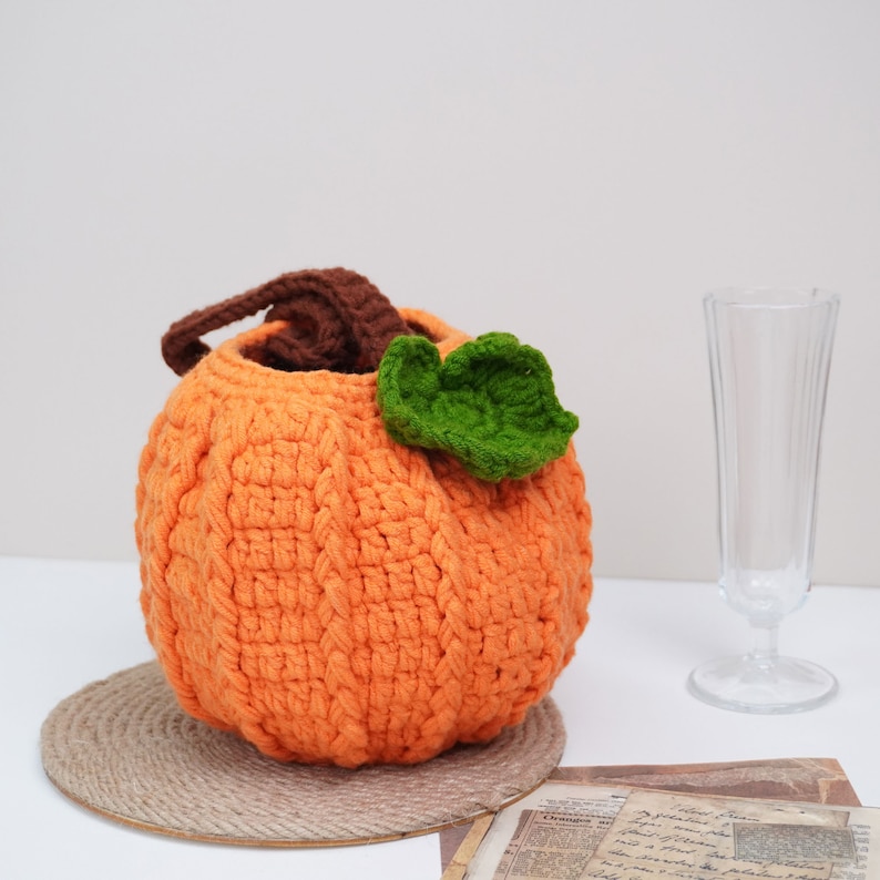May include: A crocheted orange pumpkin shaped basket with a green leaf on top. The basket is sitting on a woven round mat.