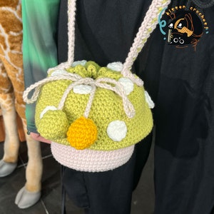 May include: A green crocheted bag with white polka dots and a yellow pom-pom. The bag has a drawstring closure and a long strap.