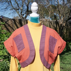 May include: A short-sleeved, open-front jacket in shades of orange and purple, with two front pockets. The jacket is layered over a long-sleeved yellow turtleneck. The garment is displayed on a mannequin.