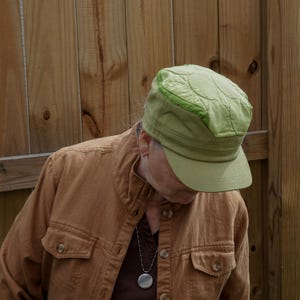 May include: A light green quilted cap with a curved brim. The cap has a structured design and a slightly darker green trim. The person is wearing a brown button-up shirt and a silver necklace with a round pendant.