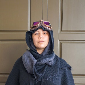 May include: A person wearing a dark gray hooded scarf and a black jacket. The person is wearing vintage-style goggles with a brown frame and purple lenses. The background is a light brown door.