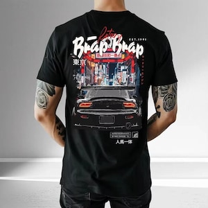 Op de afbeelding: Zwart t-shirt met een afbeelding van een sportwagen tegen een stadsachtergrond. Het ontwerp bevat de tekst "Brap Brap" in wit en rood, Japanse karakters en de woorden "EST. 1991".