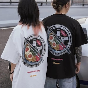 Könnte beinhalten: Zwei übergroße T-Shirts, eines weiß und eines schwarz, mit einem grafischen Design auf dem Rücken. Das Design zeigt eine stilisierte Ramen-Schüssel mit Essstäbchen, ein Autorad und den Text "DRIVER'S FAST & FOOD".
