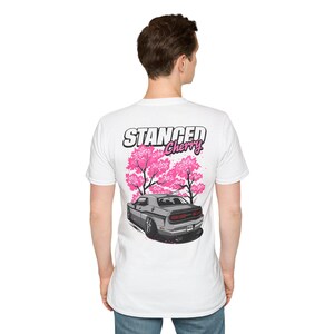 Könnte beinhalten: Weißes T-Shirt mit einer Grafik eines grauen Autos, rosa Kirschblütenbäumen und dem Text "STANCED Cherry". Das Design befindet sich auf der Rückseite. Das Shirt besteht aus einem weichen Material.