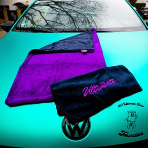 May include: Two microfiber towels, one purple and one black, with the text "The Ultimate Detailer" embroidered in pink on the black towel. The towels are on the bonnet of a turquoise car with a black Volkswagen emblem.