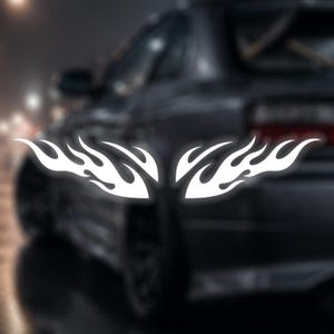 Puede incluir: Calcomanías de llamas blancas, diseñadas para ser aplicadas a un vehículo. Las calcomanías tienen un diseño de llama estilizado y simétrico. El fondo es una imagen borrosa de un coche oscuro, lo que sugiere personalización automotriz.