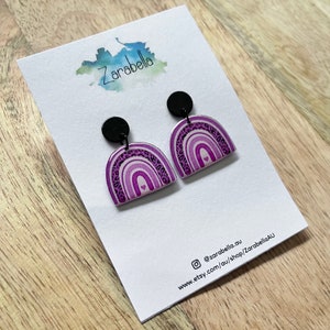 May include: A pair of purple and black rainbow earrings with a small heart in the centre of each rainbow. The earrings are made of resin and have a black metal hook.
