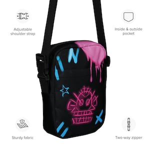 May include: Black crossbody bag with a pink and blue graffiti design. The bag has a two-way zipper, an adjustable shoulder strap, and an inside and outside pocket.