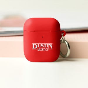 May include: Red silicone case for AirPods with a white keychain and the text "DUSTIN" printed on the front.