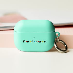 May include: A light teal AirPod case with the name "Phoebe" printed in colourful letters. The case has a silver carabiner clip attached. The case is a solid colour with a smooth finish.