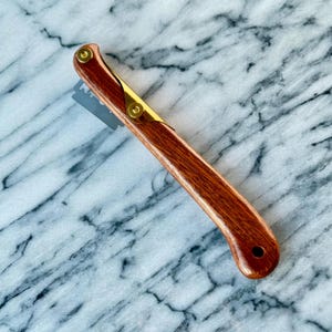 Puede incluir: Una lame de madera con un acento de latón y una cuchilla plateada. El mango es de color marrón intenso y la cuchilla está parcialmente expuesta. La lame está diseñada para marcar la masa de pan.