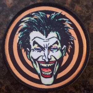 Könnte beinhalten: Ein runder, schwarz-orangener Patch mit einem weißen und grünen Bild des Jokers aus Batman. Der Joker lächelt mit einem breiten, zahnigen Grinsen. Der Hintergrund ist ein schwarz-orangenes Spiralmuster.