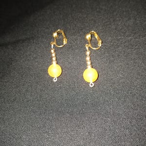 Könnte beinhalten: Ein Paar goldene Ohrclips mit einer gelben Perle, die an jedem herunterhängt.
