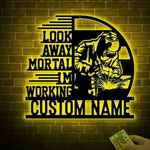 May include: A yellow illuminated metal sign with a silhouette of a welder working. The sign reads "Look away mortal I'm working CUSTOM NAME".