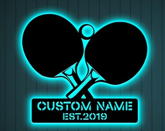 May include: A blue neon sign with two crossed ping pong paddles and a ball. The sign has a black background and the text "CUSTOM NAME EST. 2019" in white.