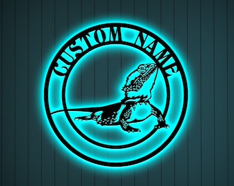 May include: A black metal cutout of a bearded dragon lizard with the words "Custom Name" around it. The cutout is illuminated with a blue light.