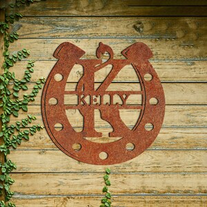 May include: A rusty metal horseshoe with the letter K in the center and the name Kelly written across the horseshoe.