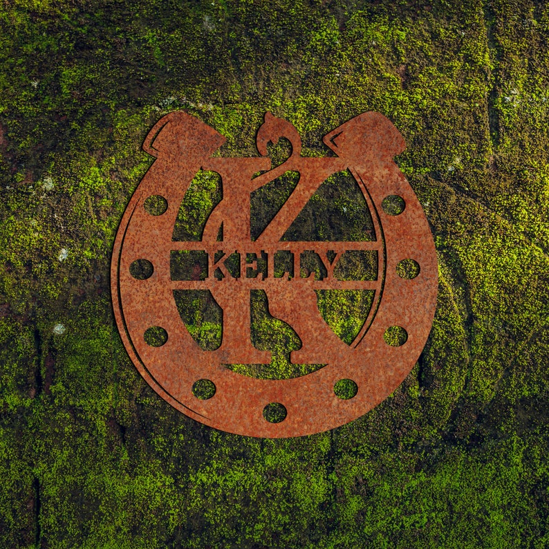 May include: A rusty metal horseshoe with the letter K in the center and the name "KELLY" written across the horseshoe. The horseshoe is mounted on a green mossy surface.