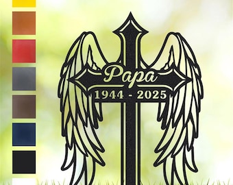 May include: Black metal memorial cross with angel wings, the word "Papa" and the years 1944-2025. The cross is set against a blurred green and yellow background, with a strip of grass at the bottom. A color palette of options is shown on the left.
