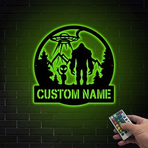 May include: A green illuminated metal sign with a silhouette of Bigfoot, an alien, and a UFO. The sign also has the text "CUSTOM NAME" on the bottom.