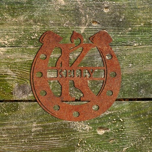 May include: Rusty metal horseshoe with the letter K and the name "Kelly" cut out. The horseshoe is on a green wooden background.