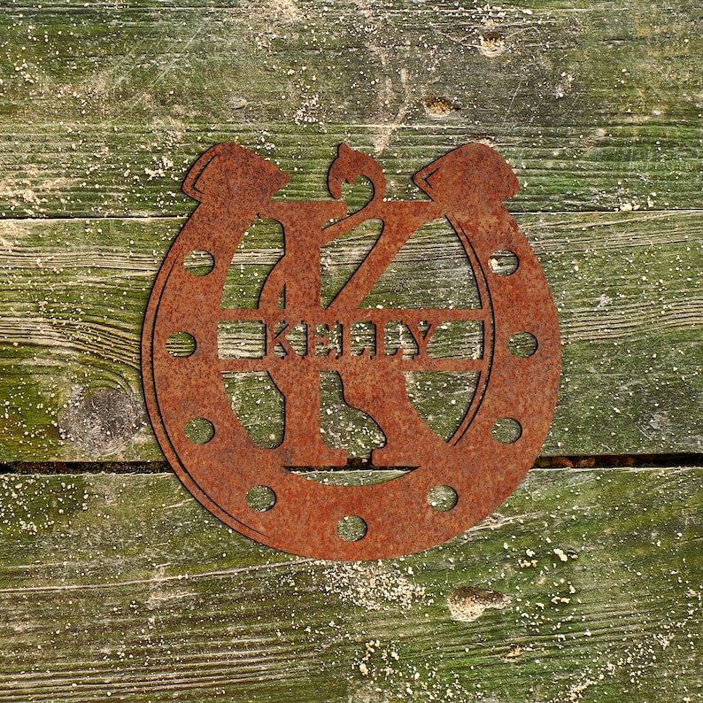 May include: Rusty metal horseshoe with the letter K and the name "Kelly" cut out. The horseshoe is on a green wooden background.