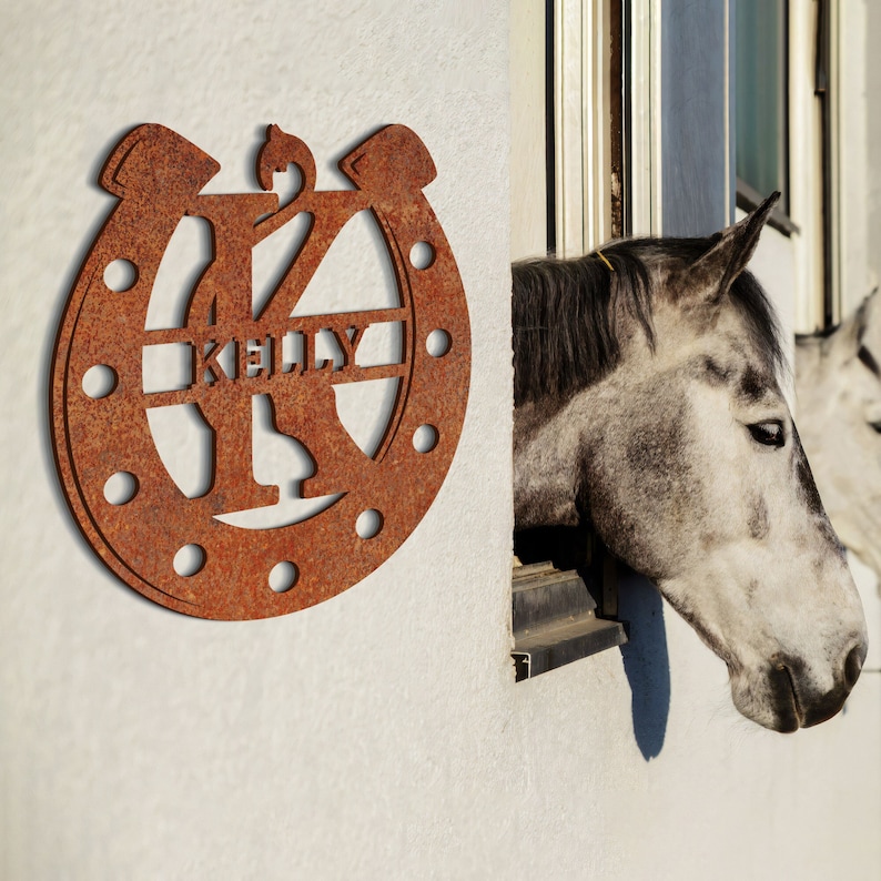 May include: A rusty metal horseshoe with the letter K and the name Kelly cut out. The horseshoe is mounted on a white wall. A gray horse's head is peeking out of a window behind the horseshoe.