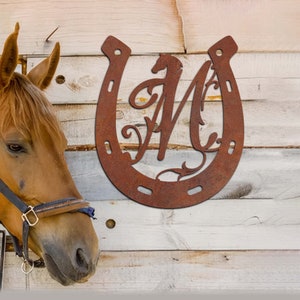 May include: A rusty brown metal horseshoe with a decorative letter M inside. The horseshoe is on a light wood background.