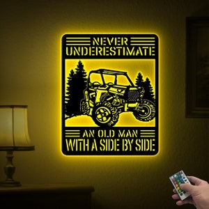 May include: A yellow neon sign with the text "Never underestimate an old man with a side by side."  The sign features a silhouette of a side-by-side vehicle in a forest setting.
