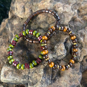 May include: Three wooden bead bracelets in shades of brown, green, red, and gold. The bracelets feature a variety of bead shapes and sizes, with some accented by gold-colored metal spacers. The bracelets are displayed on a textured stone surface.