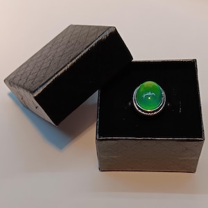 May include: A silver ring with a large, oval, green gemstone is displayed in an open black ring box. The gemstone has a bright, glossy appearance. The ring box is textured with a diamond pattern.