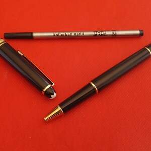 May include: A black Montblanc rollerball pen with gold accents and a white star logo. The pen is next to a silver rollerball refill with the text "Rollerball Refill" and "MONT BLANC" printed on it.