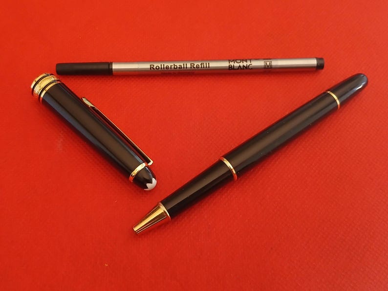 May include: A black Montblanc rollerball pen with gold accents and a white star logo. The pen is next to a silver rollerball refill with the text "Rollerball Refill" and "MONT BLANC" printed on it.