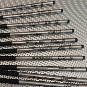 Könnte beinhalten: Eine Sammlung von neun silberfarbenen Rollerball-Stiftminen mit schwarzen Kunststoffkappen. Jede Mine ist mit schwarzem Text bedruckt, der Rollerball Refill, Montblanc, Made in Germany sowie ein quadratisches M-Symbol zeigt.