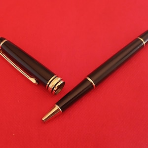 Könnte beinhalten: Ein schwarzer und goldener Stift mit Kappe und Clip. Der Stift liegt auf einer roten Oberfläche.