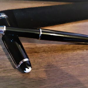 May include: A black rollerball pen with silver detailing and a white emblem on the cap. The pen rests on a wooden surface beside a black pen case. The pen's cap is open, showing the pen's writing tip.