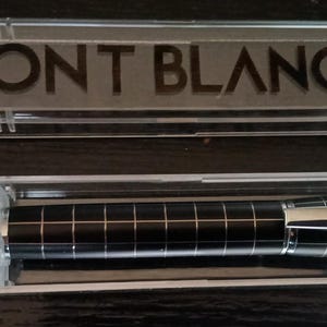 May include: A Montblanc ballpoint pen in a clear plastic case. The pen has a black checkered pattern with silver accents and a silver tip. The case lid displays the Montblanc logo and brand name.
