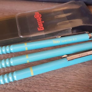 May include: Three turquoise mechanical pencils with silver accents and the word "Writing" printed on the side of one pencil. The pencils are in a clear plastic case with a black backing that has the word "Writing" printed on it in red.