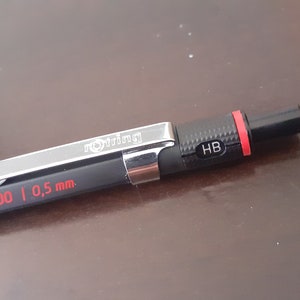 Peut inclure: Un crayon mécanique noir avec un clip argenté et des accents rouges. Le crayon est marqué "rotring 300 | 0,5 mm" et "HB".