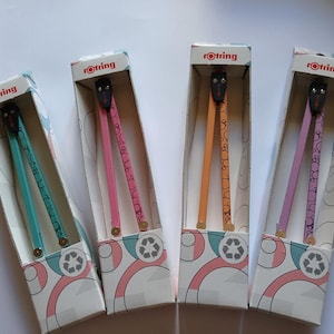 Può includere: Quattro compassi Rotring in scatole. Ogni compasso ha una testa nera e bracci colorati in verde acqua, rosa, arancione e viola. Le scatole sono bianche con un design rosa, verde acqua e pesca e un simbolo di riciclaggio.