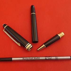 May include: A black rollerball pen with gold accents and a black rollerball refill. The pen has the words "MONT BLANC" and "M" printed on it. The refill has the words "Rollerball Refill" printed on it.