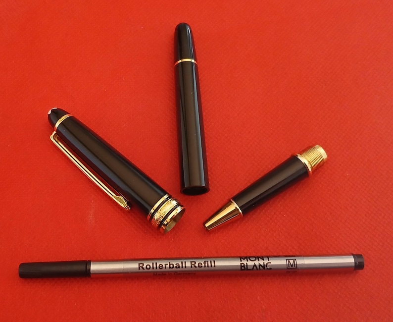 May include: A black rollerball pen with gold accents and a black rollerball refill. The pen has the words "MONT BLANC" and "M" printed on it. The refill has the words "Rollerball Refill" printed on it.