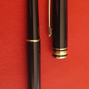 May include: Two black pens with gold accents. The pen on the left has a black cap and a gold band near the tip. The pen on the right has a white star on the cap and a gold band near the tip.