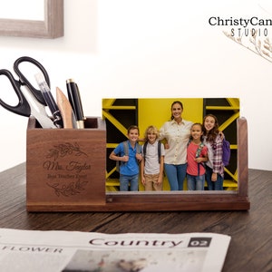 May include: Wooden desk organiser with a photo frame insert. The photo frame shows a teacher and students. The organiser has a compartment for pens and pencils and a small drawer. The organiser is engraved with the text "Mrs. Taylor Best Teacher Ever!"