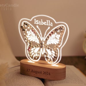 Puede incluir: Una luz nocturna con forma de mariposa blanca con una base de madera. La mariposa tiene un diseño floral y está decorada con pequeñas cuentas y pedrería. La base tiene el nombre "Isabella" y la fecha "17 de agosto de 2024" grabada.