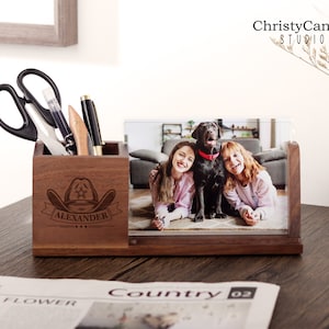 以下が含まれることがあります： 写真立てとペンやその他のオフィス用品を入れるためのコンパートメントが付いた木製デスクオーガナイザー。写真立てには、2 人の人と犬の写真が飾られています。オーガナイザーには、「Alexander」というテキストとカウボーイハットのグラフィックが刻印されています。